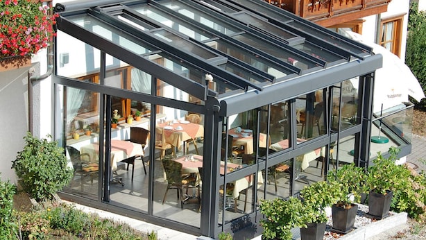 Une terrasse moderne, fermée par des parois de verre, avec des tables et des chaises, entourée de plantes en pot et d’un bâtiment doté d’un balcon.