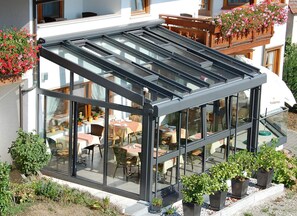 Een moderne, glazen patio met tafels en stoelen, omringd door potplanten en een gebouw met een balkon.