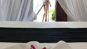 A bed with a white cover and pink flowers, a window with sheer curtains, and a view of a person outside. - Savera Beach Houses (Jambiani)
