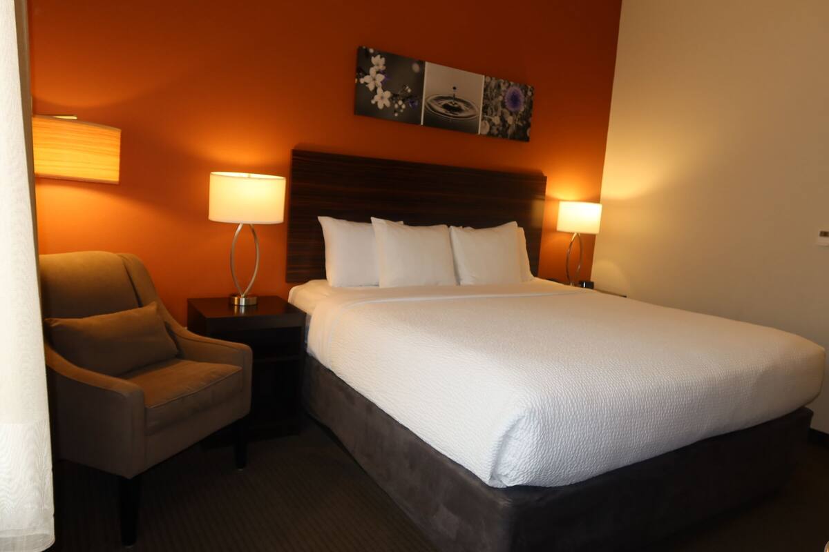Quarto de hotel com cama, cadeira, mesa de cabeceira e duas luminárias fixadas na parede.