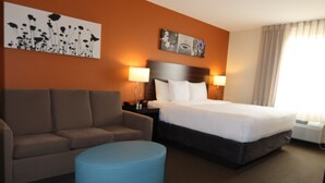 A hotel room with a bed, a sofa, a blue ottoman, a nightstand, and wall art. - Sleep Inn & Suites Oregon - Madison (Oregon)