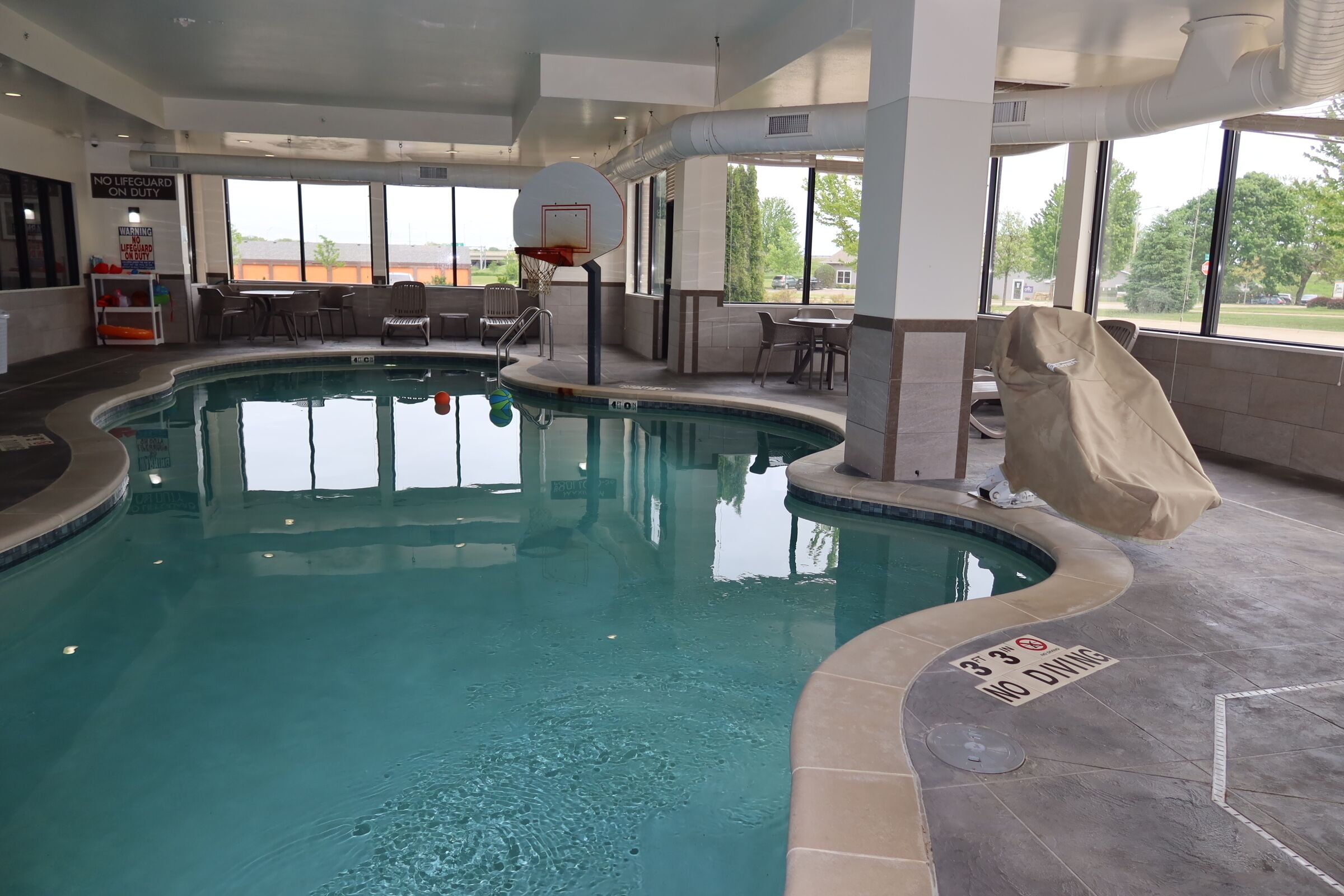 An indoor swimming pool with a no diving sign, a basketball hoop, and a covered seating area.