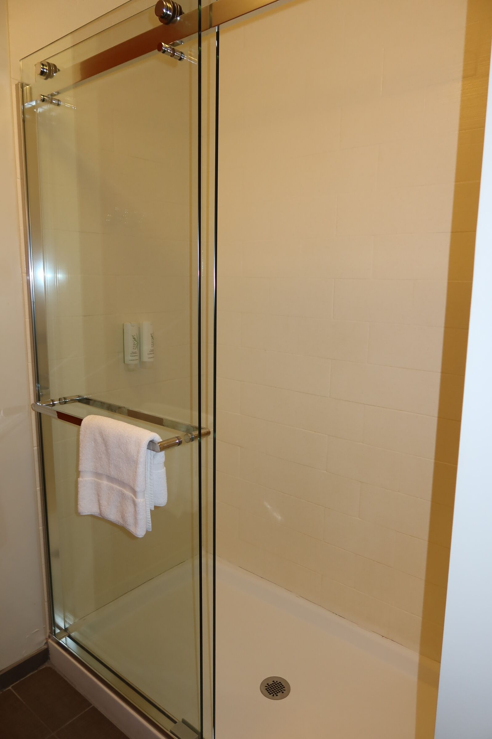 A glass-enclosed shower with a towel rack, a white tiled wall, and a visible drain.