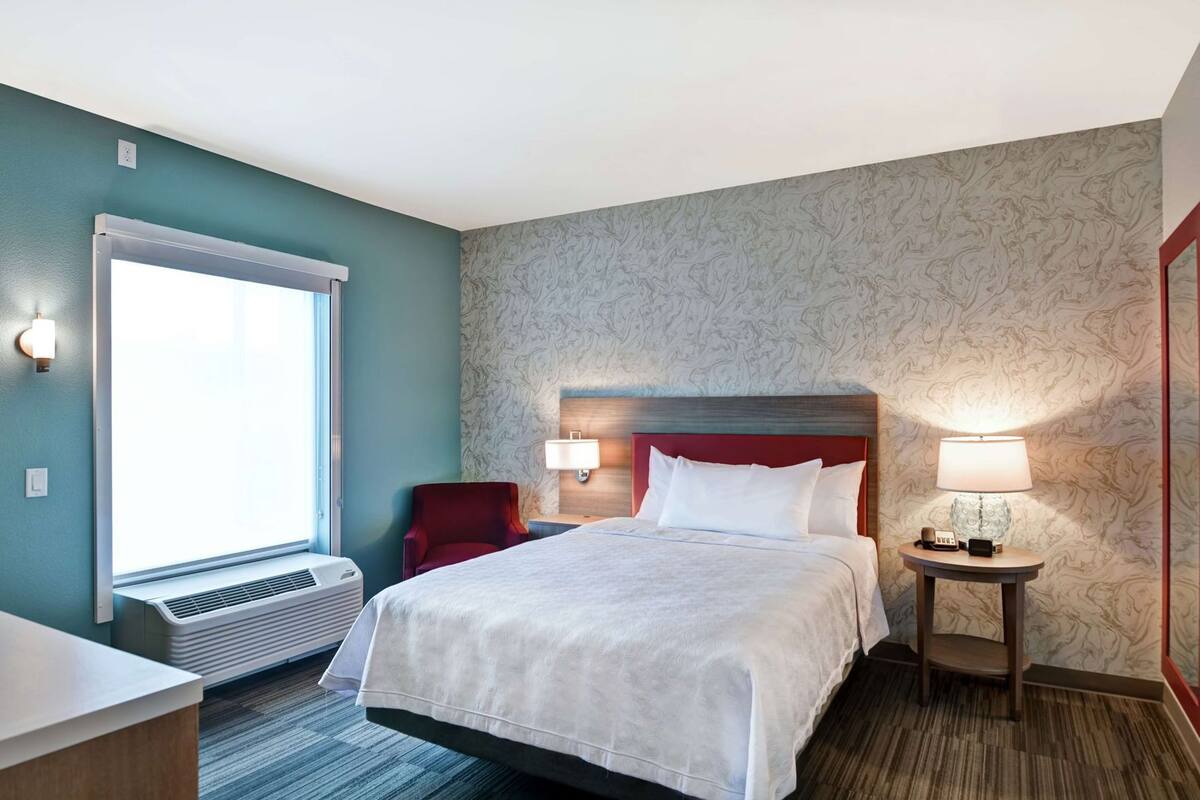 Habitación de hotel con cama, una silla, escritorio, lámpara y ventana.