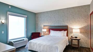 A hotel room with a bed, a desk, a chair, a nightstand with a lamp, and a window with an air conditioner.
