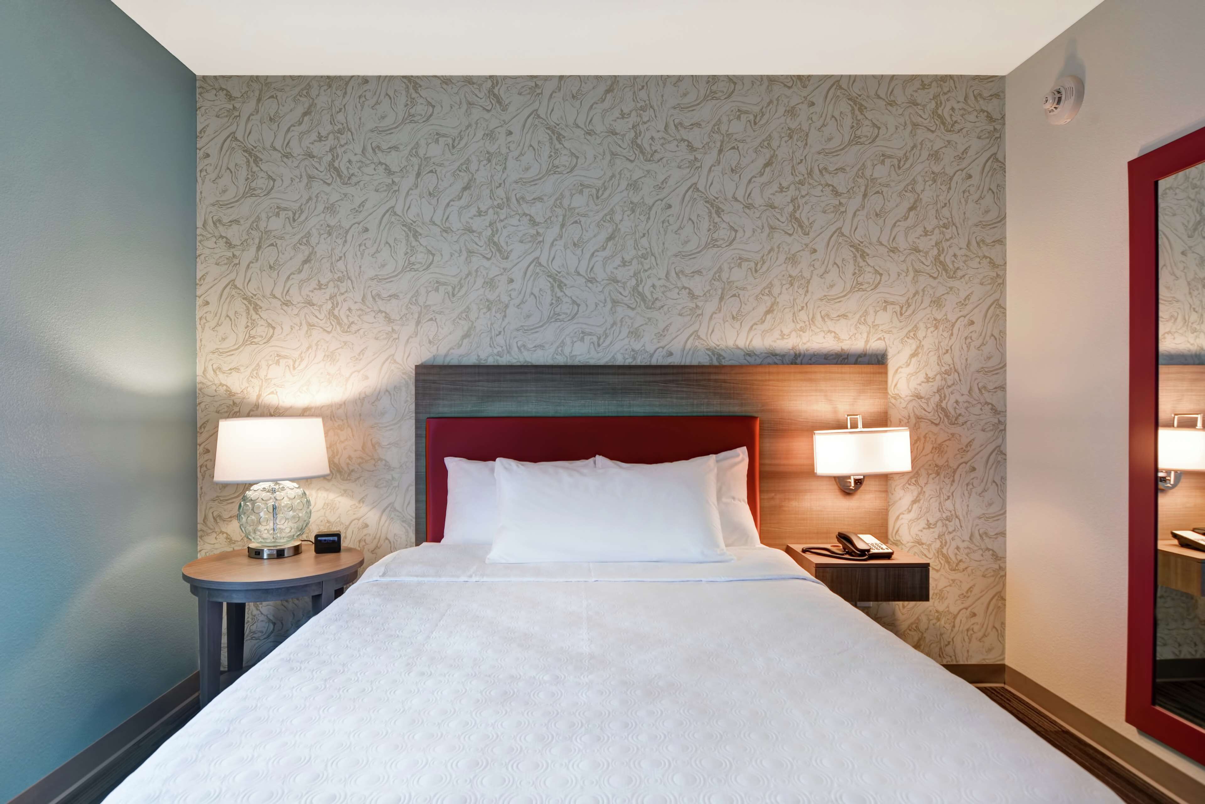 A hotel room with a large bed, two bedside tables with lamps, a red headboard, and patterned wallpaper.