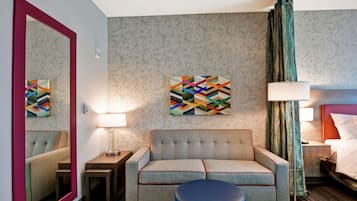 A hotel room with a sofa, a round ottoman, a mirror, a wall art piece, a lamp, and a bed.
