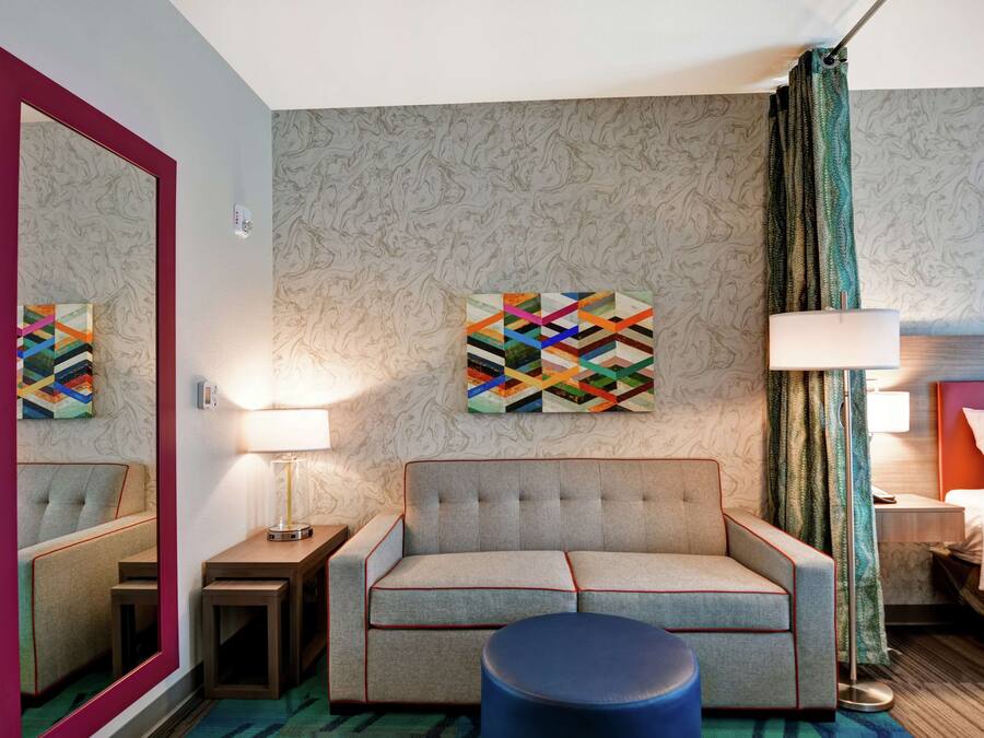 A hotel room with a sofa, a round ottoman, a mirror, a wall art piece, a lamp, and a bed.