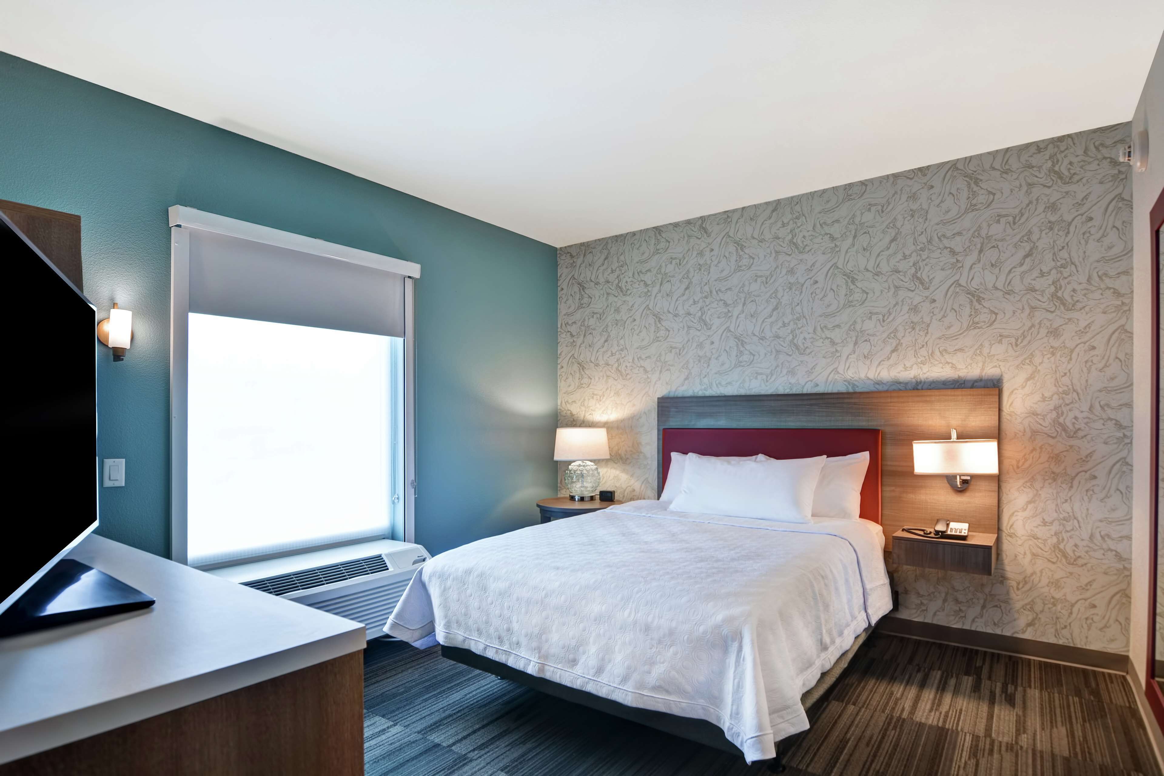 A hotel room with a bed, bedside tables with lamps, a desk with a laptop, and a window with blinds.