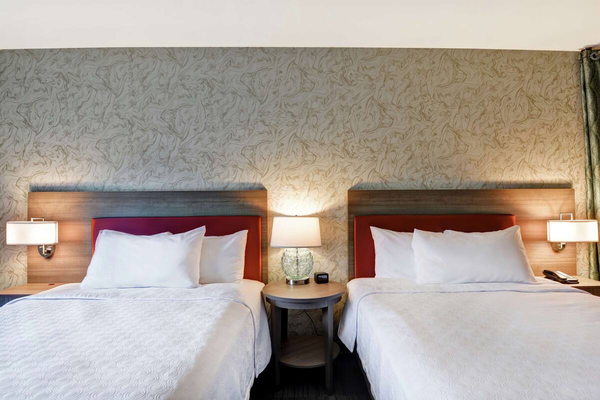 Habitación de hotel con dos camas, cada una con cabecero rojo, una mesita redonda con lámpara y un teléfono en la pared.