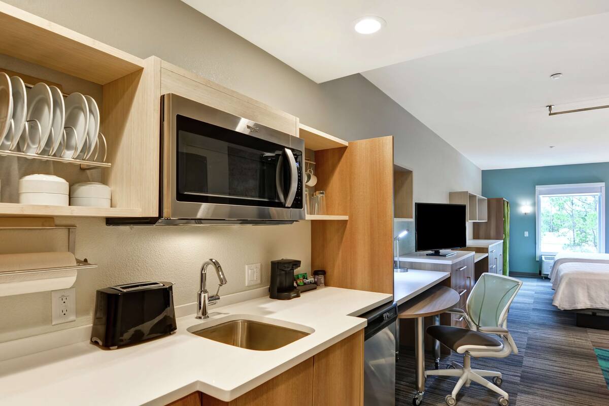 Una cocina moderna con microondas, tostadora y lavabo. Hay un comedor con una silla y un televisor. La habitación tiene una cama y una ventana con vistas a árboles.