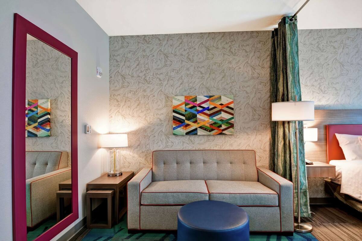 Una habitación de hotel con sofá, espejo, mesita de noche, lámpara, cama y una obra de arte colorida en la pared.