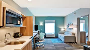 A modern hotel room with a kitchenette, a sofa, a desk, and a large window providing natural light.