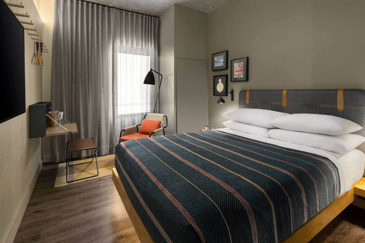 Una habitación de hotel moderna con una cama grande, un escritorio con una silla, una lámpara de pie y una ventana con cortinas.