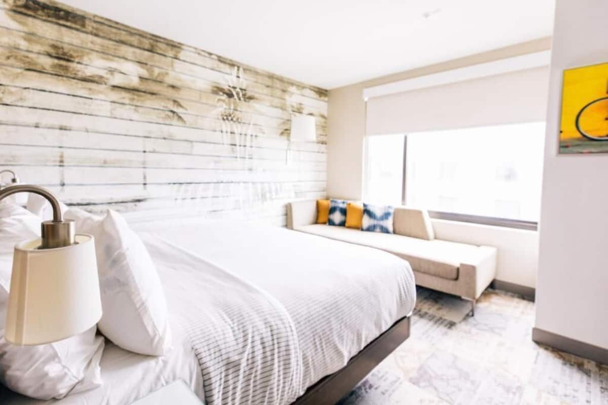 A modern bedroom with a wooden headboard, a large bed with white bedding, a bedside table with lamps, and a window with a view.