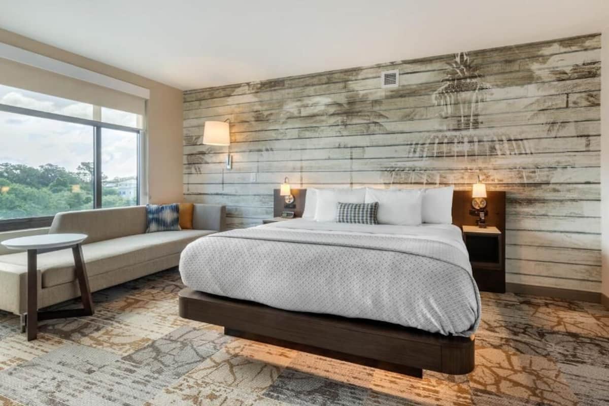 A modern hotel room with a large bed, a sofa, a small table, and a view of the outdoors.