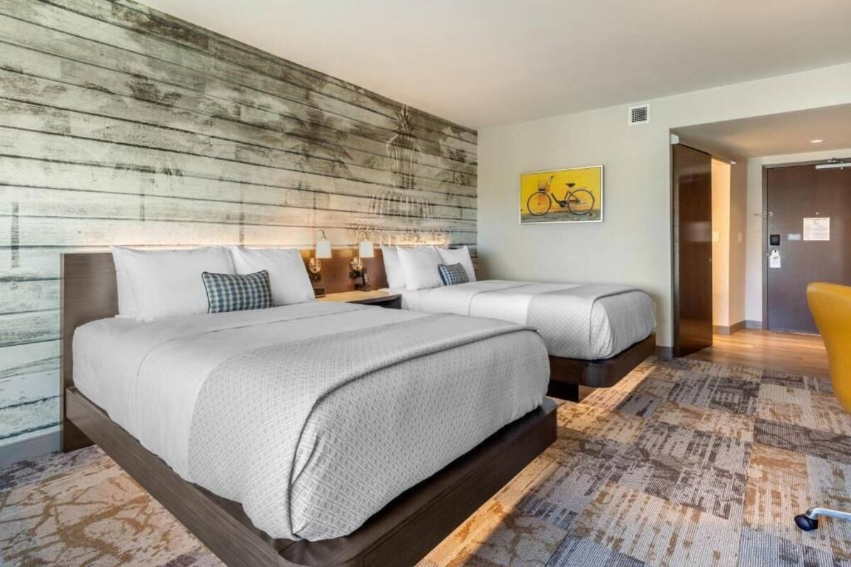 A hotel room with two beds, a wooden accent wall, a patterned rug, and a bicycle artwork on the wall.