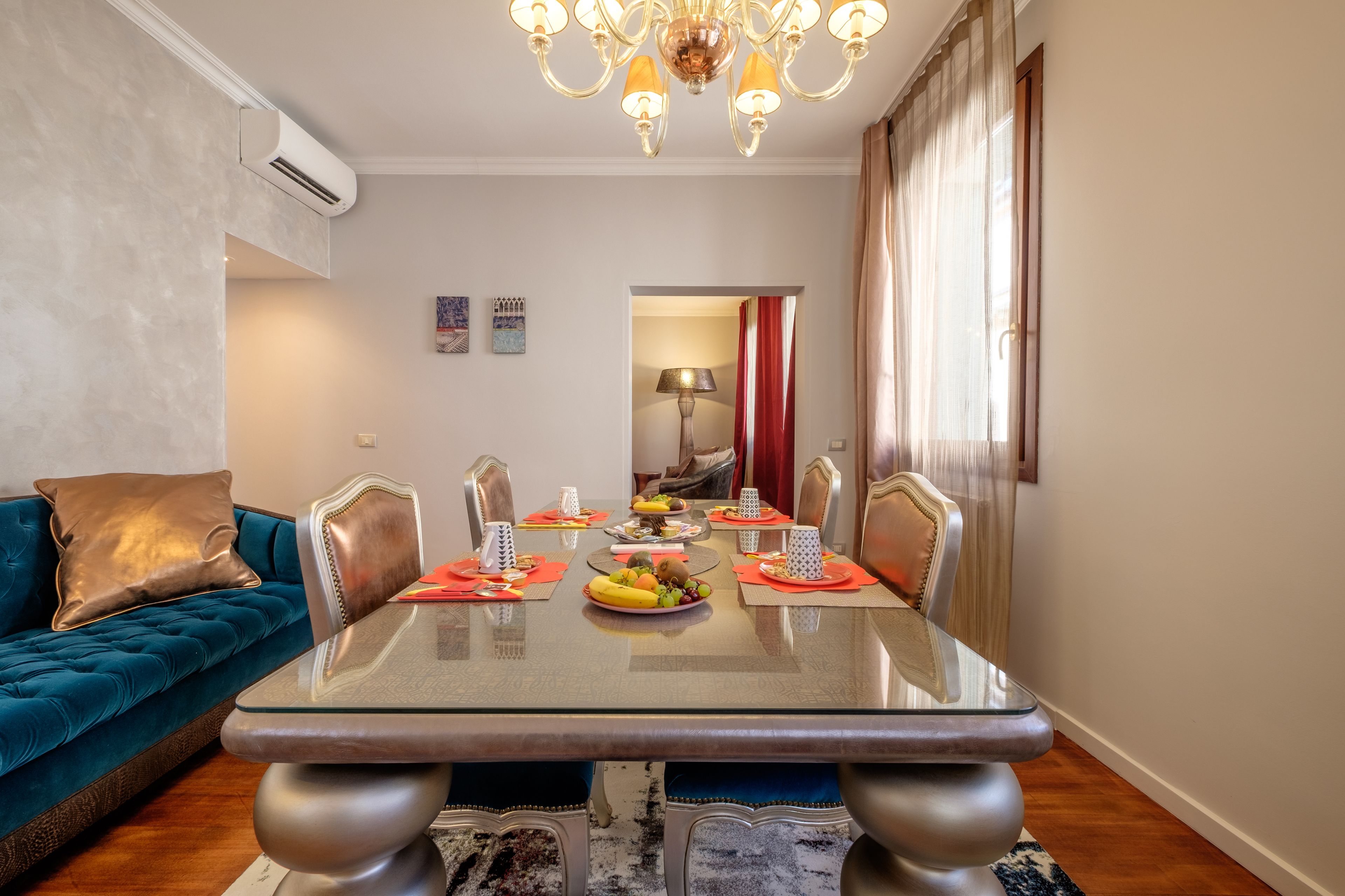 Un comedor con una mesa de vidrio, individuales rojos y un frutero. Hay un sofá azul con un cojín marrón y una araña colgante.