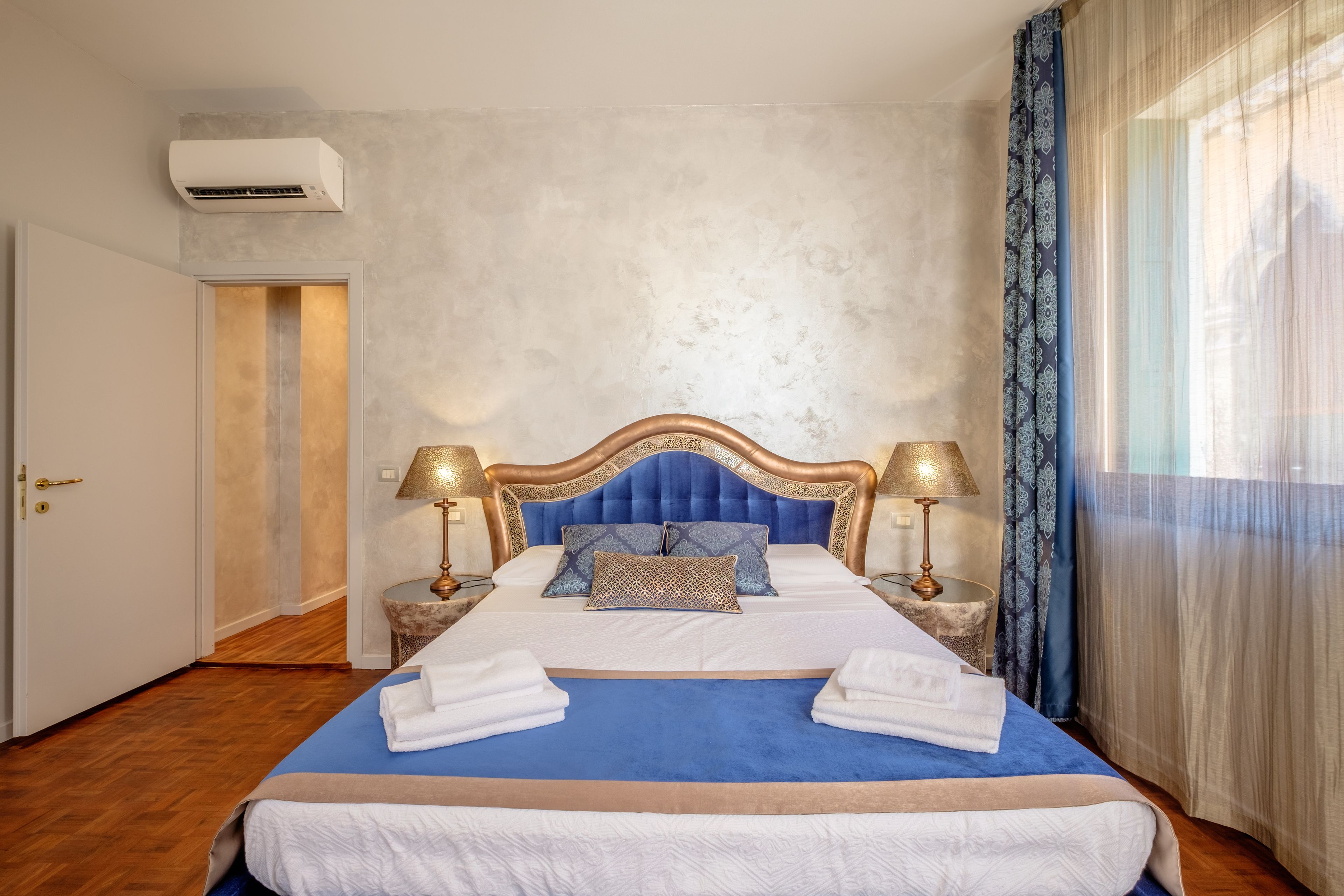 A neatly made bed with a blue bedspread, white linens, and two decorative pillows. There are two bedside lamps with gold bases and lampshades. The room has a textured wall and a large window with sheer curtains.