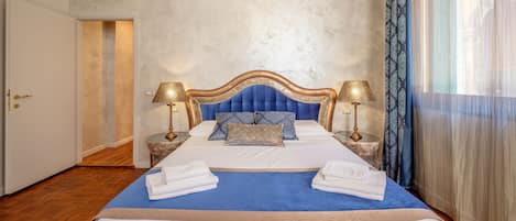 A neatly made bed with a blue bedspread, white linens, and two decorative pillows. There are two bedside lamps with gold bases and lampshades. The room has a textured wall and a large window with sheer curtains.