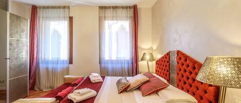 Une chambre d’hôtel avec un grand lit, une tête de lit rouge, une lampe de chevet et deux fenêtres ornées de rideaux légers.