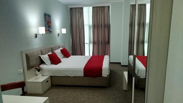 A hotel room with a bed, bedside tables, a nightstand, a mirror, and a window with curtains. - Green Tower Hotel (Tbilisi)