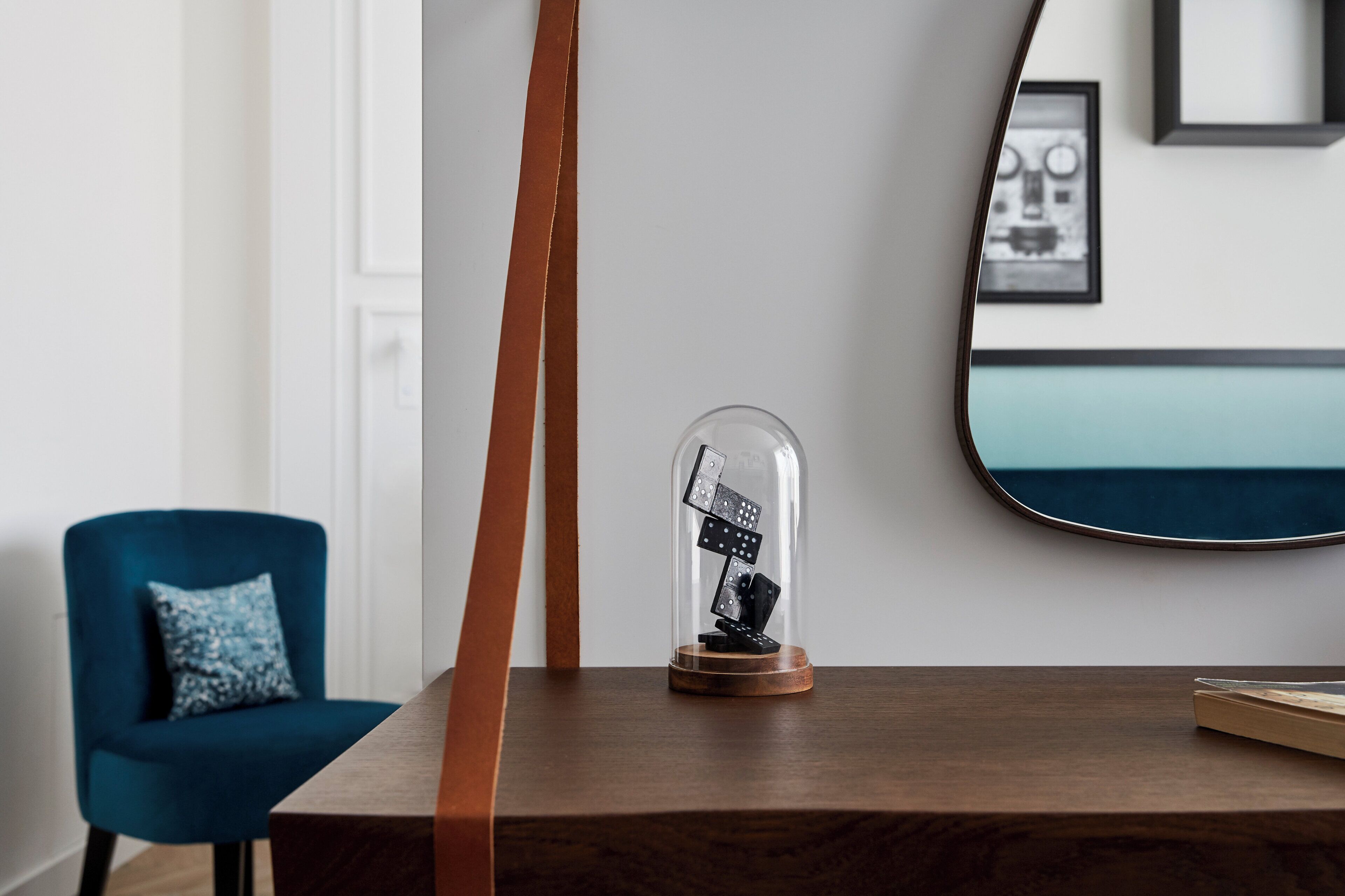 A wooden table with a glass dome containing black objects, a blue chair with a patterned pillow, and a mirror on the wall.