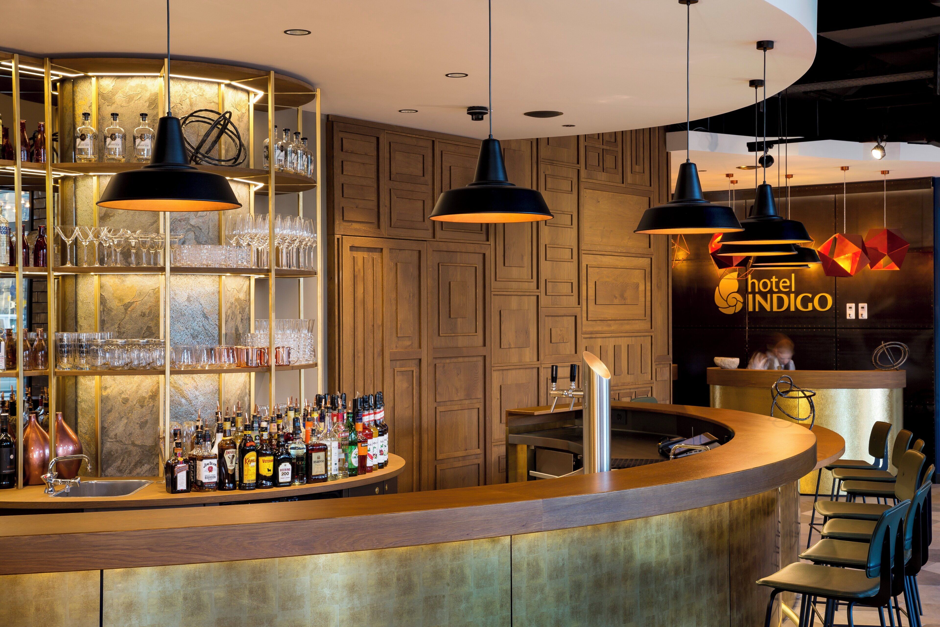 A bar area with a curved counter, shelves with bottles, glasses, and a sign for 'hotel INDIGO'.