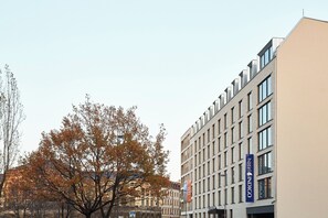 Un moderno edificio de hotel con un llamativo letrero 'Hotel Indigo', múltiples ventanas y un árbol con hojas otoñales en primer plano.