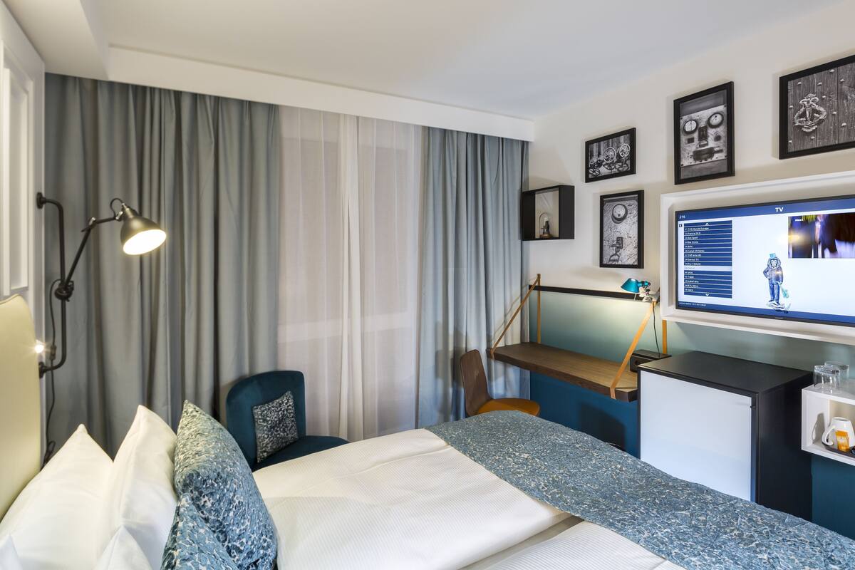 A hotel room with a bed, a desk, a chair, a TV, and a window with curtains.