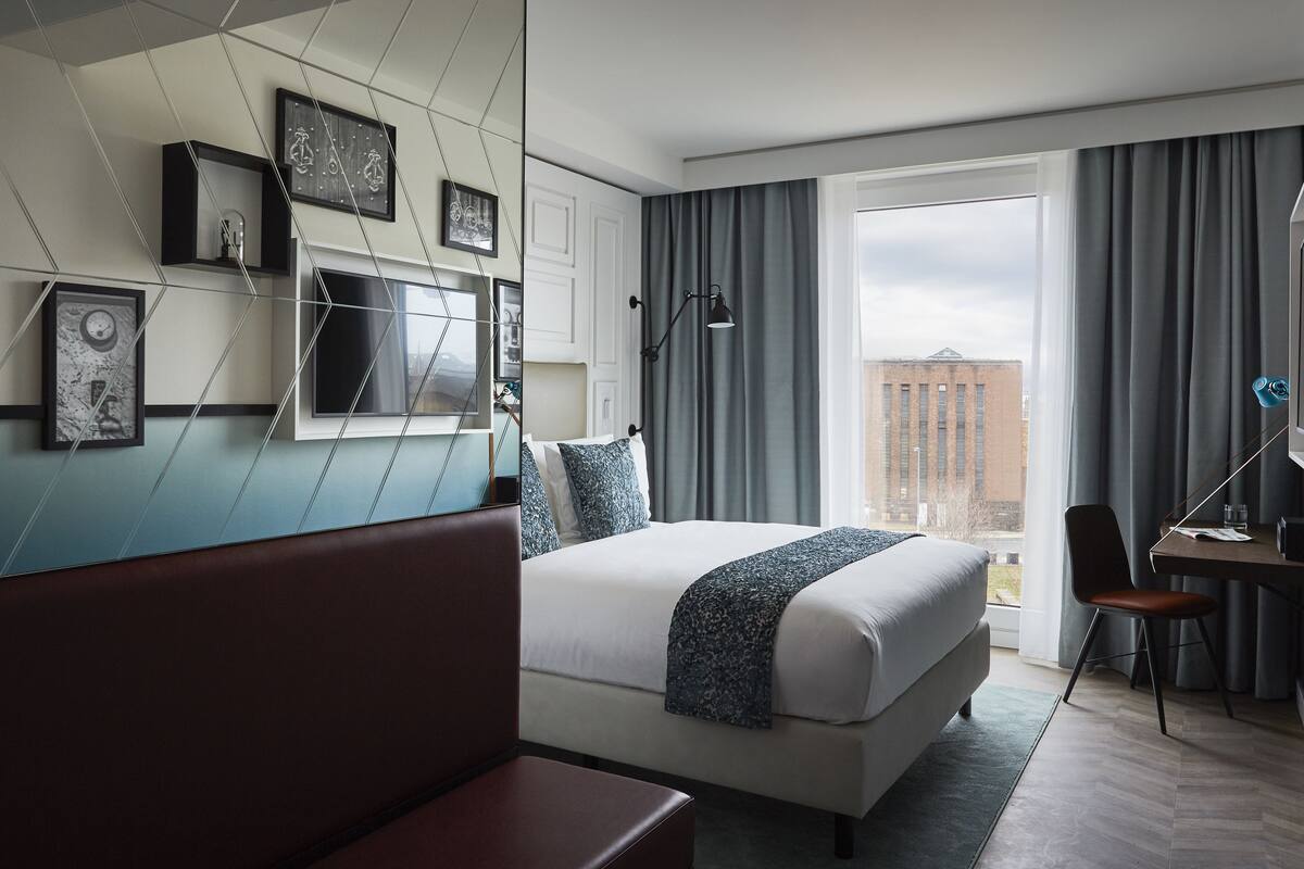 A modern hotel room with a large bed, a desk with a chair, and a city view through the window.