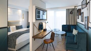A modern hotel room with a bed, a desk with a chair, a sofa, and a TV mounted on the wall.