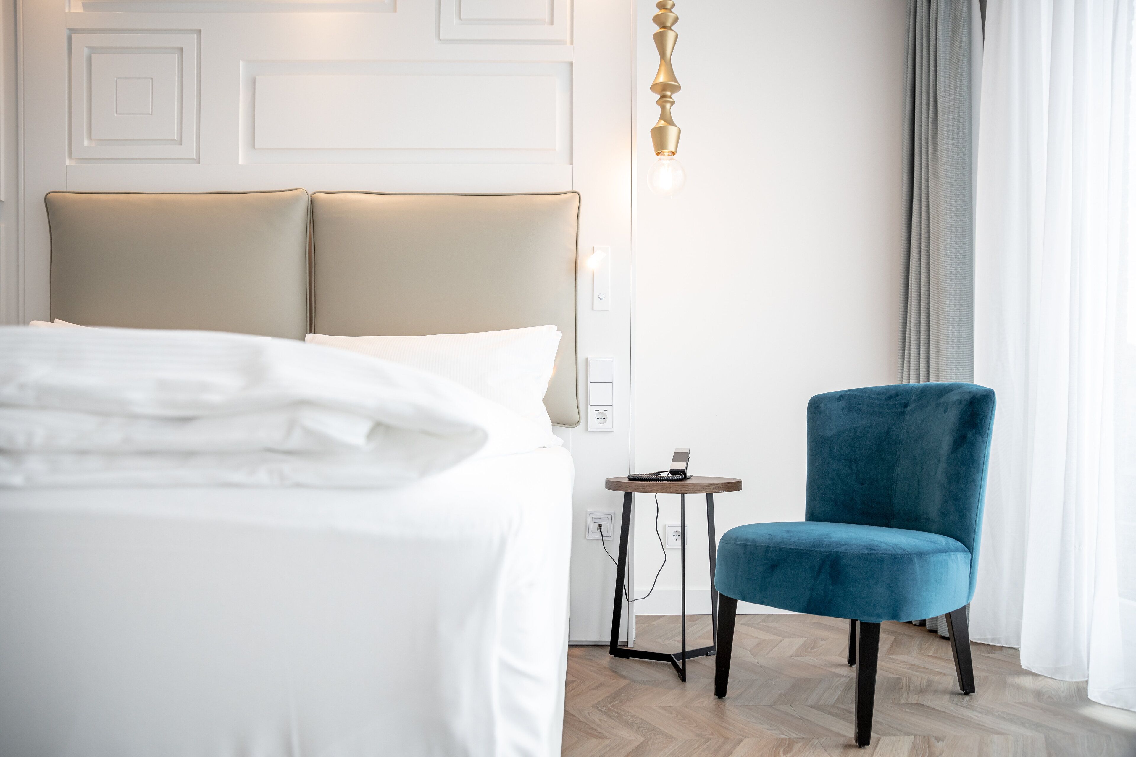 A hotel room with a bed, a small table with a phone, a blue chair, and a wall-mounted light fixture.