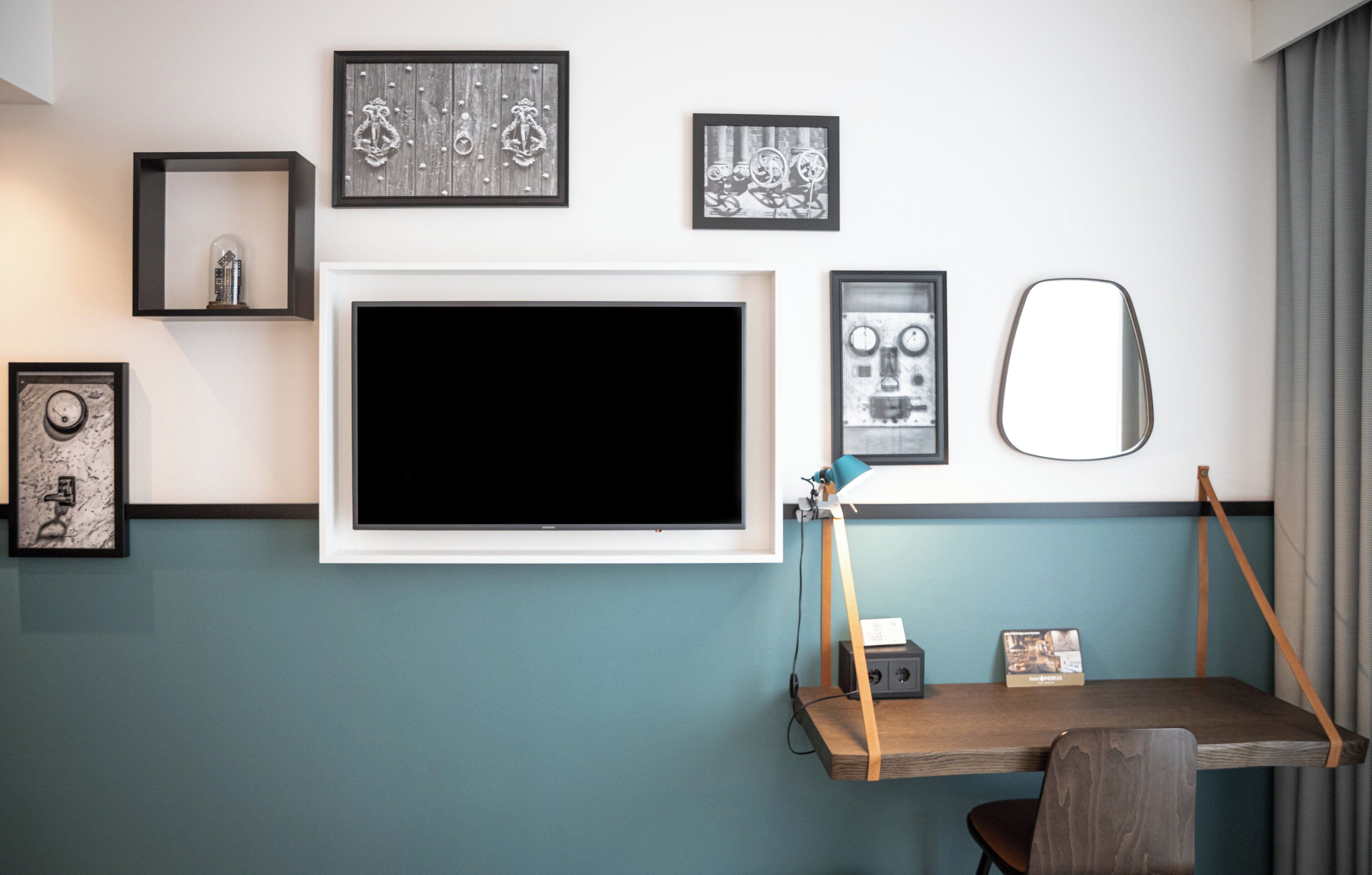 A room with a mounted TV, framed artwork, a desk with a chair, and a mirror.