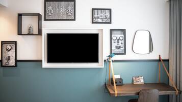 A room with a mounted TV, framed artwork, a desk with a chair, and a mirror.