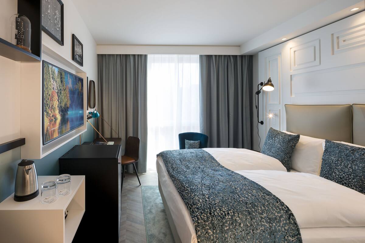 A hotel room with a bed, a desk with a coffee maker, a chair, a desk lamp, and a painting on the wall.