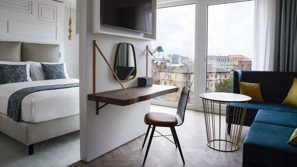 A modern hotel room with a bed, a desk, a chair, a TV, and a sofa.