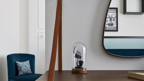 A wooden table with a glass dome containing black and white dominoes, a blue chair with a patterned pillow, and a mirror on the wall.