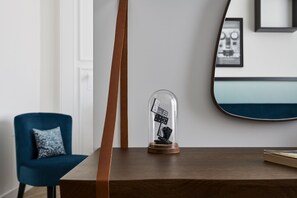 Una mesa de madera con una cúpula de vidrio que contiene dominó blanco y negro, una silla azul con un cojín estampado, y un espejo en la pared.