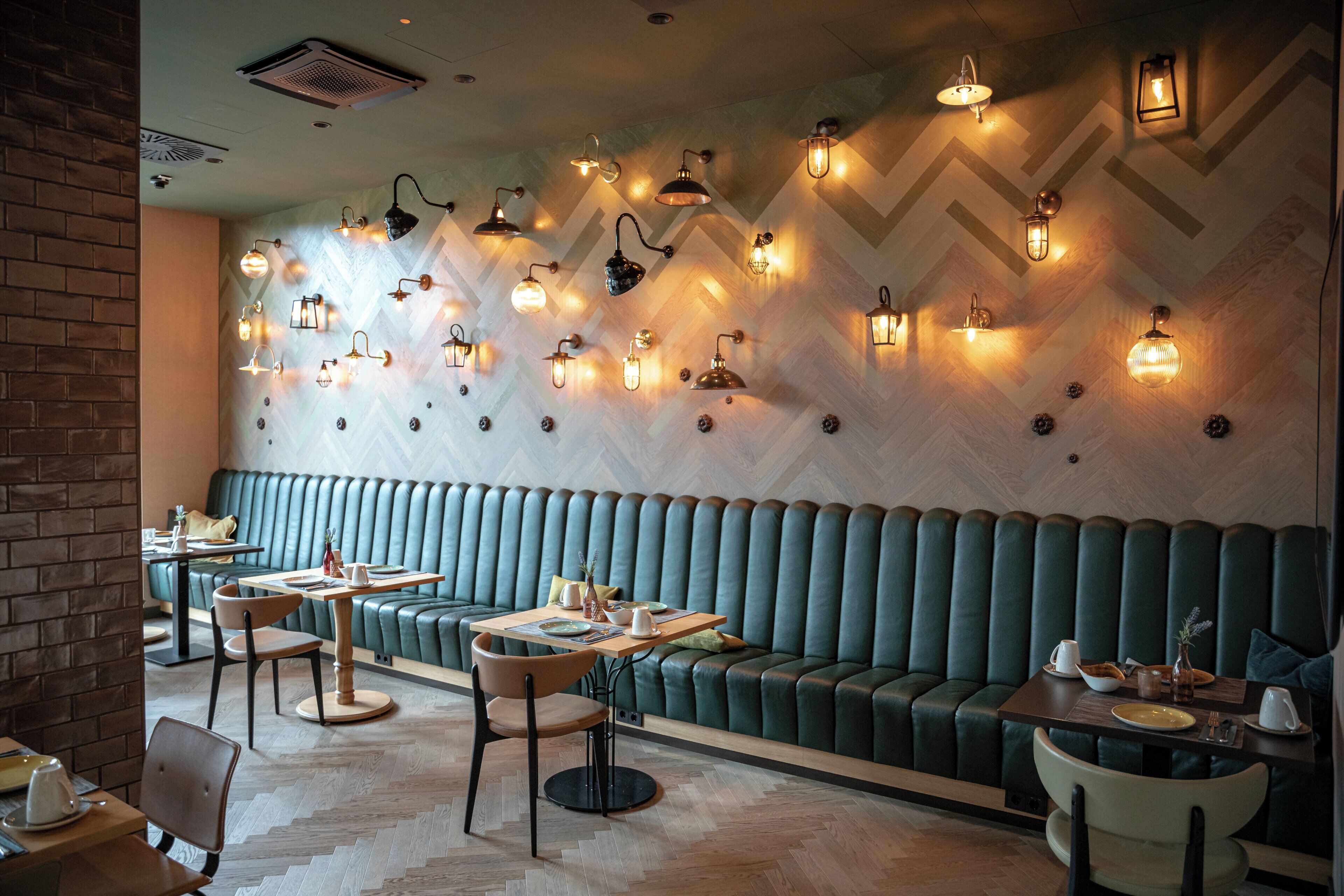A restaurant interior with a herringbone patterned wall, green upholstered booths, and multiple tables with chairs.