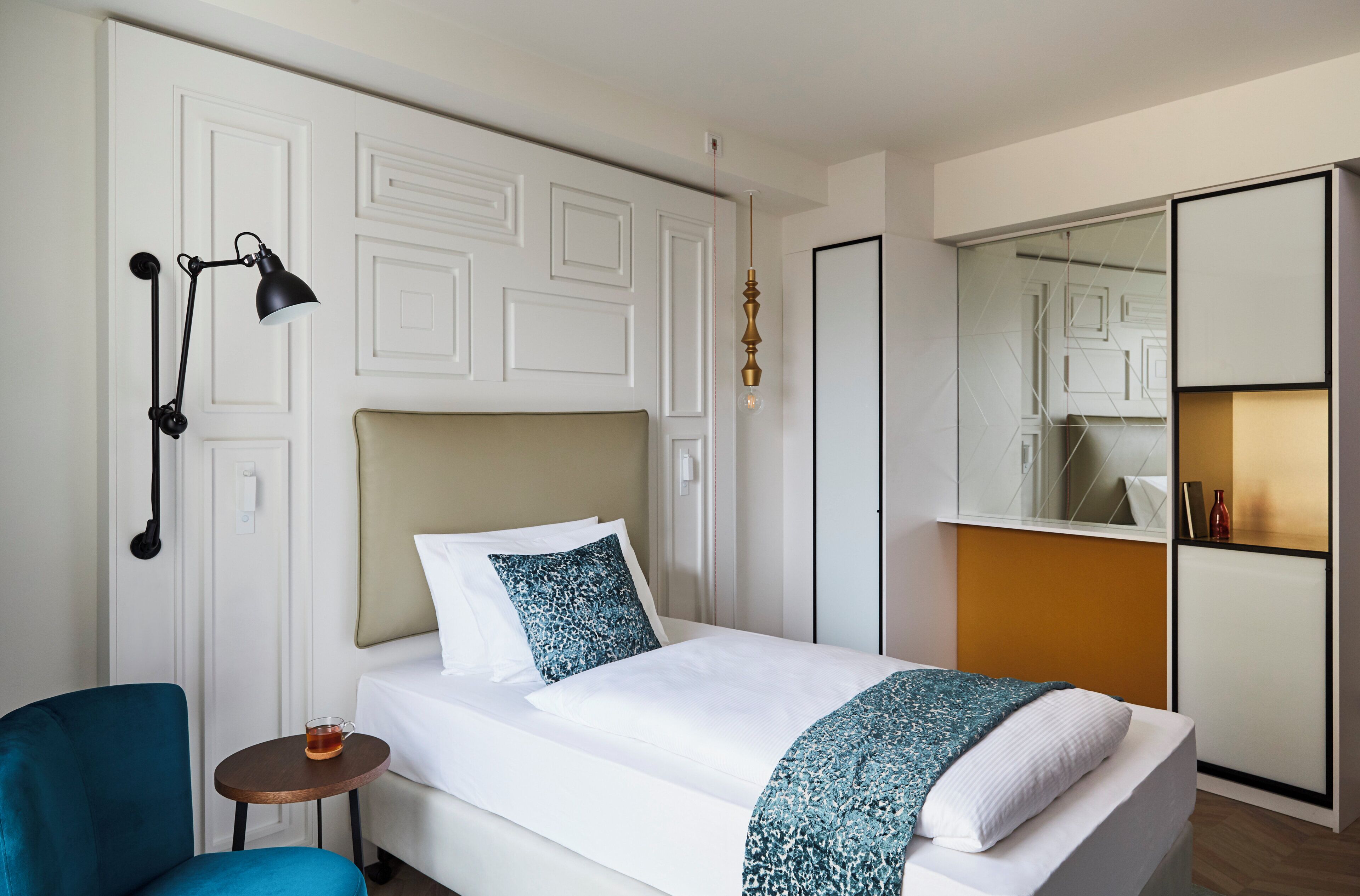 A hotel room with a bed, bedside table, chair, and a mirror with a shelf.