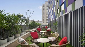 An outdoor seating area with wicker furniture and red cushions, surrounded by potted plants and a striped building facade. - ibis Kolkata Rajarhat Hotel (Barasat)