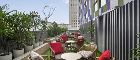 An outdoor seating area with wicker furniture and red cushions, surrounded by potted plants and a striped building facade.