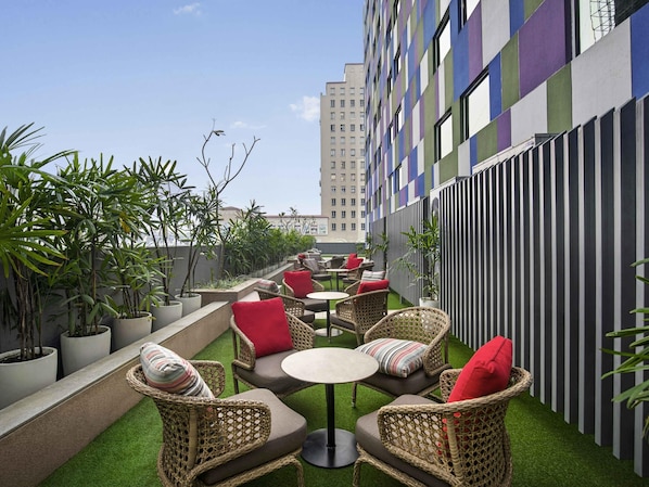 An outdoor seating area with wicker furniture and red cushions, surrounded by potted plants and a striped building facade.