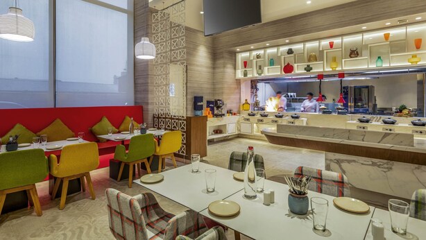 A modern restaurant with a central bar area, dining tables with chairs, and a kitchen in the background. - ibis Kolkata Rajarhat Hotel (Barasat)