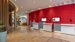 A modern reception area with a red geometric wall, wooden reception desks, and a curved ceiling design. - ibis Kolkata Rajarhat Hotel (Barasat)