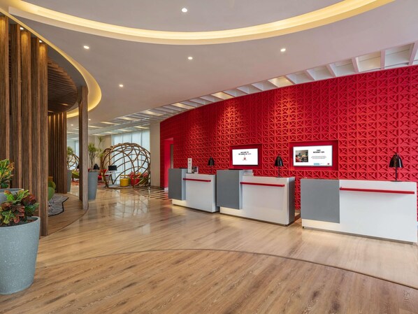 A modern reception area with a red geometric wall, wooden reception desks, and a curved ceiling design. - ibis Kolkata Rajarhat Hotel (Barasat)