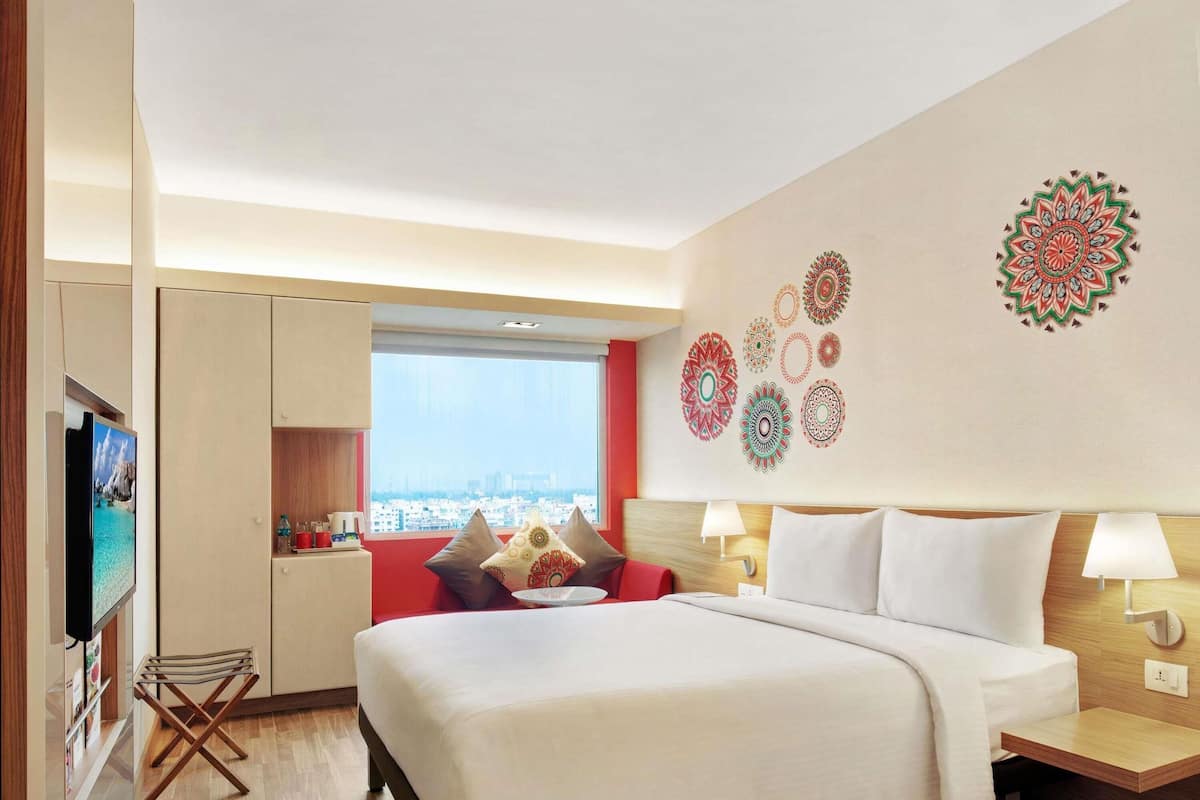 Habitación de hotel con una cama, un televisor, una silla roja, una mesita de noche de madera y una pared con diseños decorativos.
