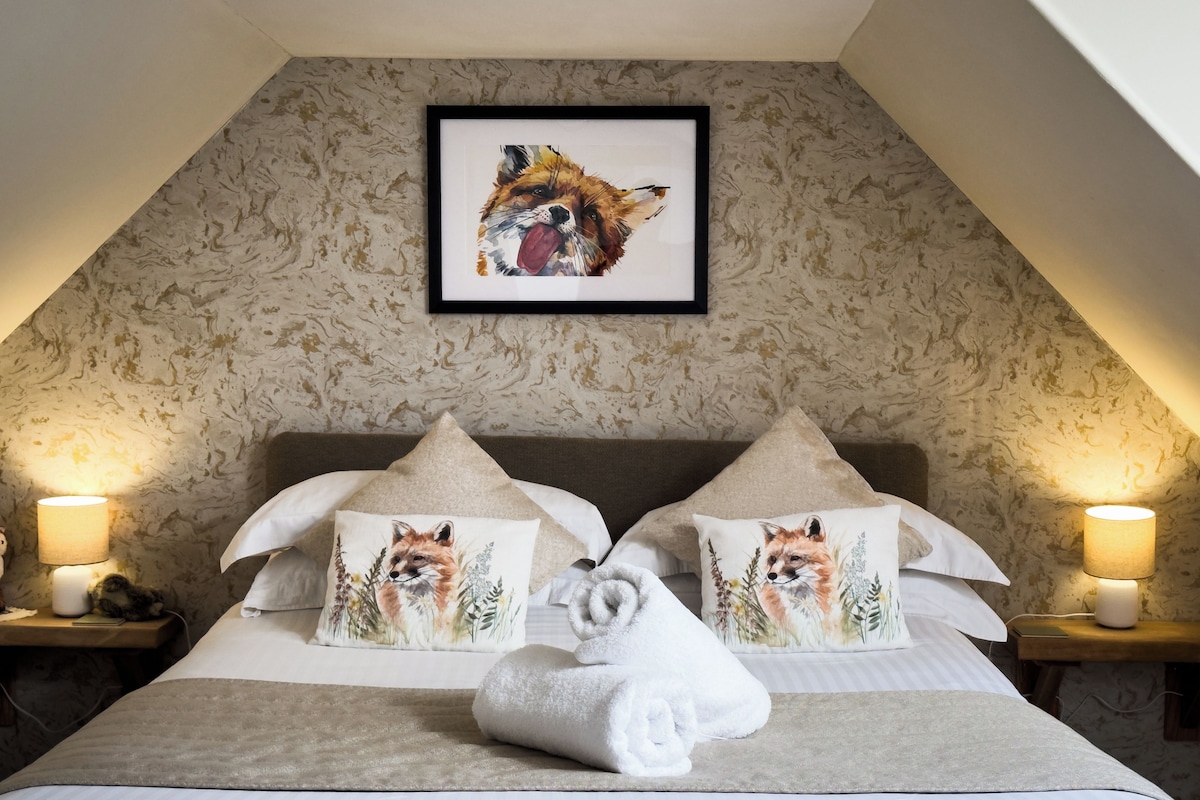 A neatly made bed with two pillows featuring fox illustrations, a framed artwork above, and a lamp on each side.