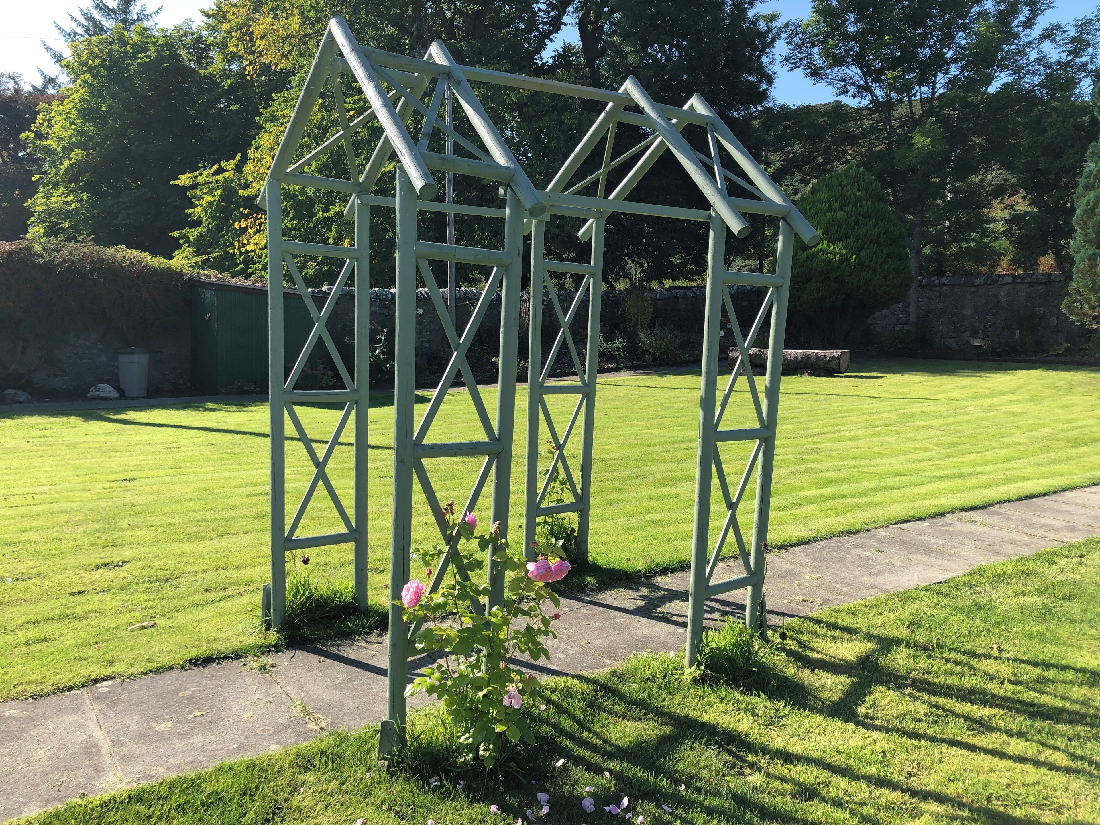 A green metal structure with a triangular roof, surrounded by a well-maintained lawn and a stone wall in the background.